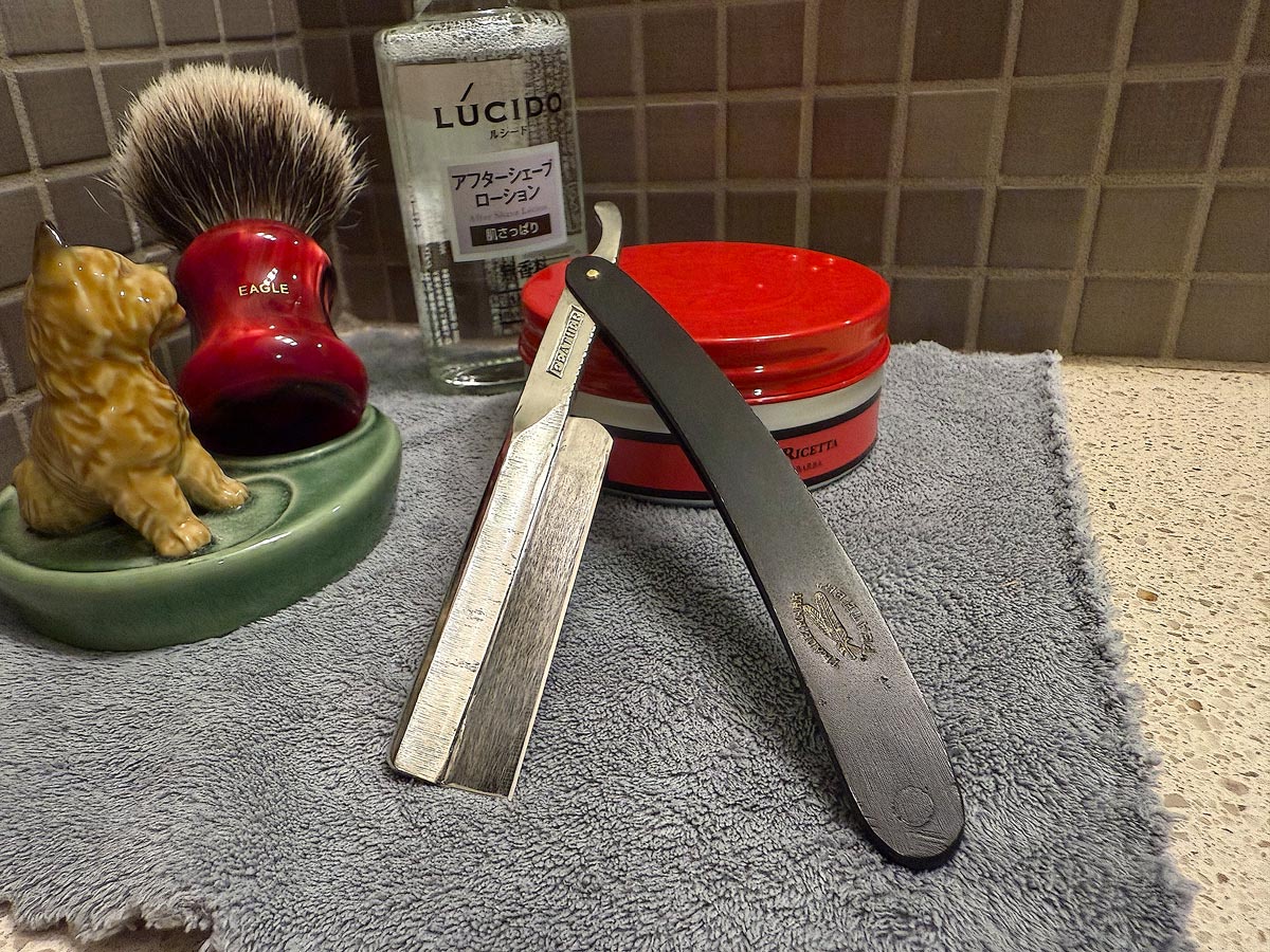 On a gray microfiber towel a straight razor with blade open about 45 degrees leans next to a bottle of aftershave and a jar of shave soap. A shave brush rests bristles-up in a green Wade ceramic pipe rest that features a brown Cairn Terrier.
