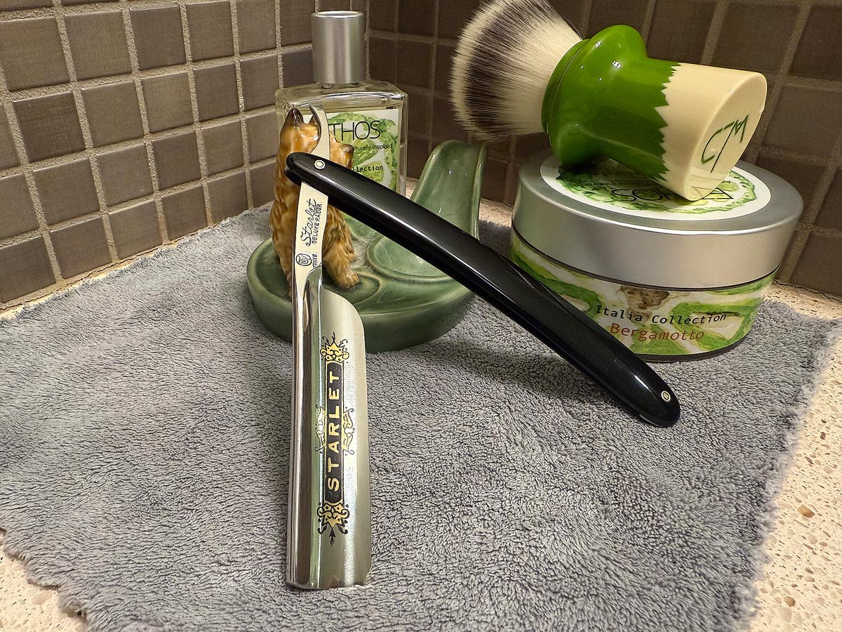 On a gray microfiber towel a straight razor with blade open about 45 degrees leans next to a bottle of aftershave and a jar of shave soap. A shave brush rests bristles-up in a green Wade ceramic pipe rest that features a brown Cairn Terrier.