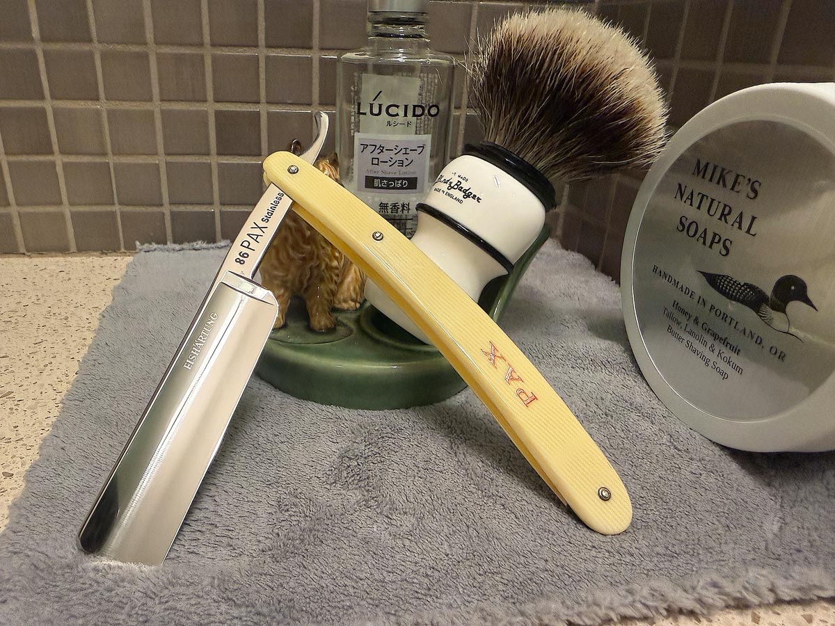 On a gray microfiber towel a straight razor with blade open about 45 degrees leans next to a bottle of aftershave and a jar of shave soap. A shave brush rests bristles-up in a green Wade ceramic pipe rest that features a brown Cairn Terrier.