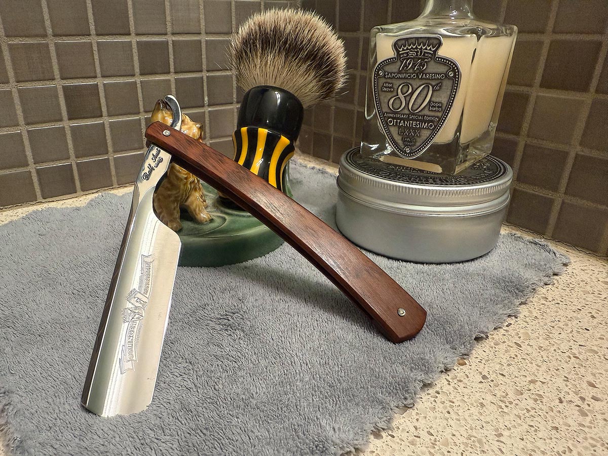 On a gray microfiber towel a straight razor with blade open about 45 degrees leans next to a bottle of aftershave and a jar of shave soap. A shave brush rests bristles-up in a green Wade ceramic pipe rest that features a brown Cairn Terrier.