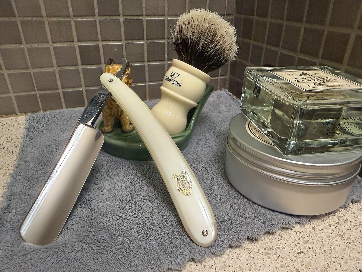 On a gray microfiber towel a straight razor with blade open about 45 degrees leans next to a bottle of aftershave and a jar of shave soap. A shave brush rests bristles-up in a green Wade ceramic pipe rest that features a brown Cairn Terrier.