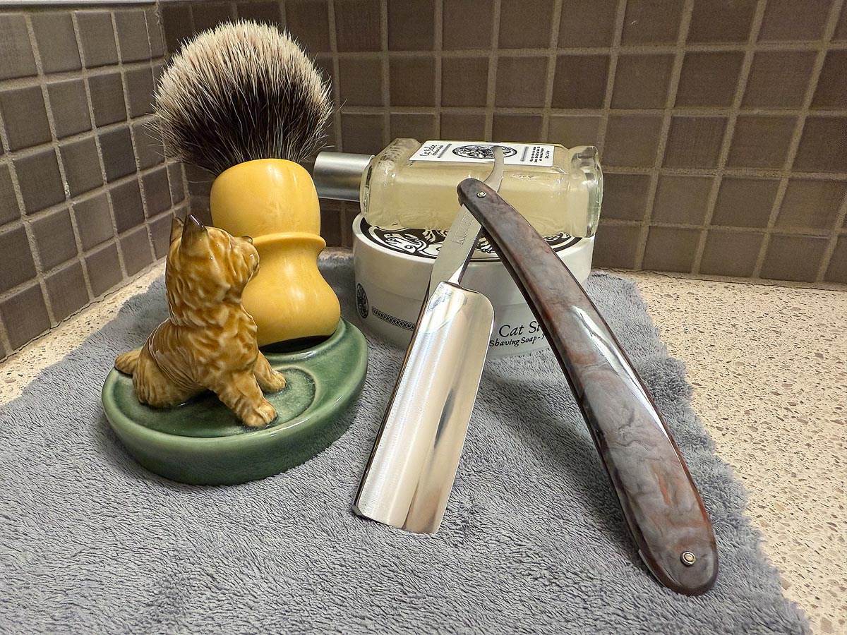 On a gray microfiber towel a straight razor with blade open about 45 degrees leans next to a bottle of aftershave and a jar of shave soap. A shave brush rests bristles-up in a green Wade ceramic pipe rest that features a brown Cairn Terrier.