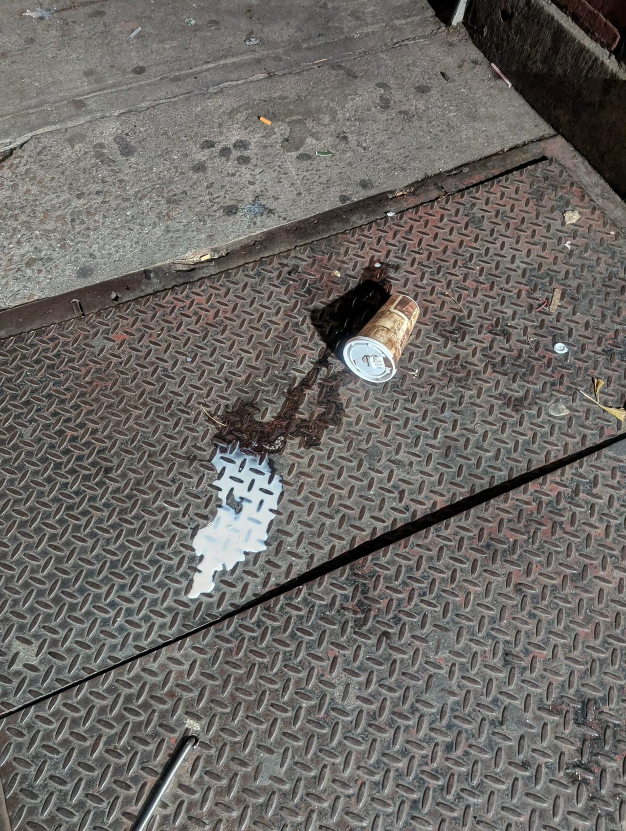 a spilled coffee cup on a metal basement door on the sidewalk, a milky liquid spilled from its end and threatening to pour through the doors' crack