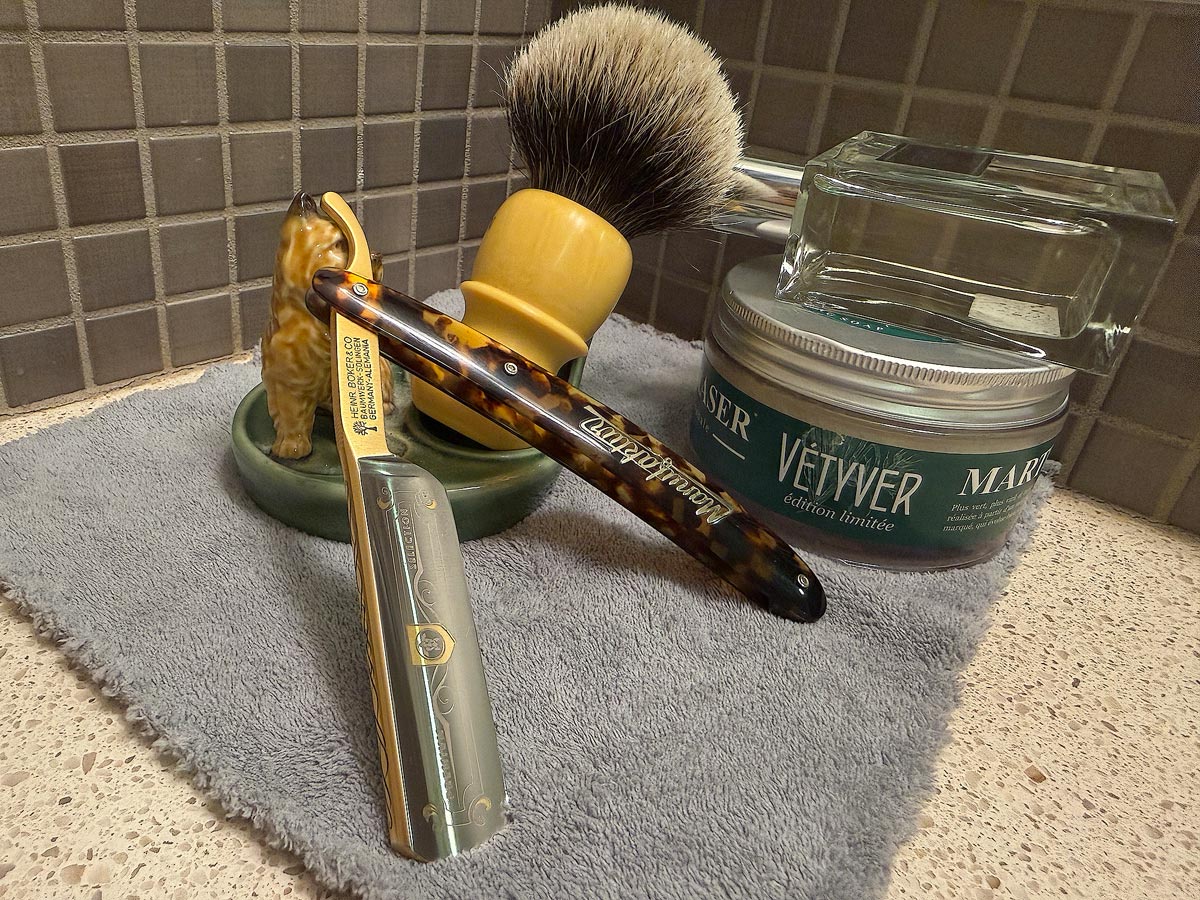 On a gray microfiber towel a straight razor with blade open about 45 degrees leans next to a bottle of aftershave and a jar of shave soap. A shave brush rests bristles-up in a green Wade ceramic pipe rest that features a brown Cairn Terrier.