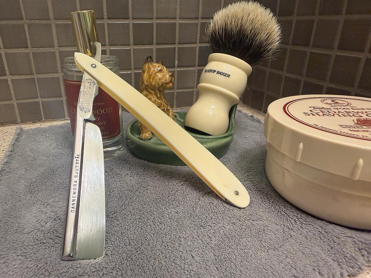 On a gray microfiber towel a straight razor with blade open about 45 degrees leans next to a bottle of aftershave and a jar of shave soap. A shave brush rests bristles-up in a green Wade ceramic pipe rest that features a brown Cairn Terrier.