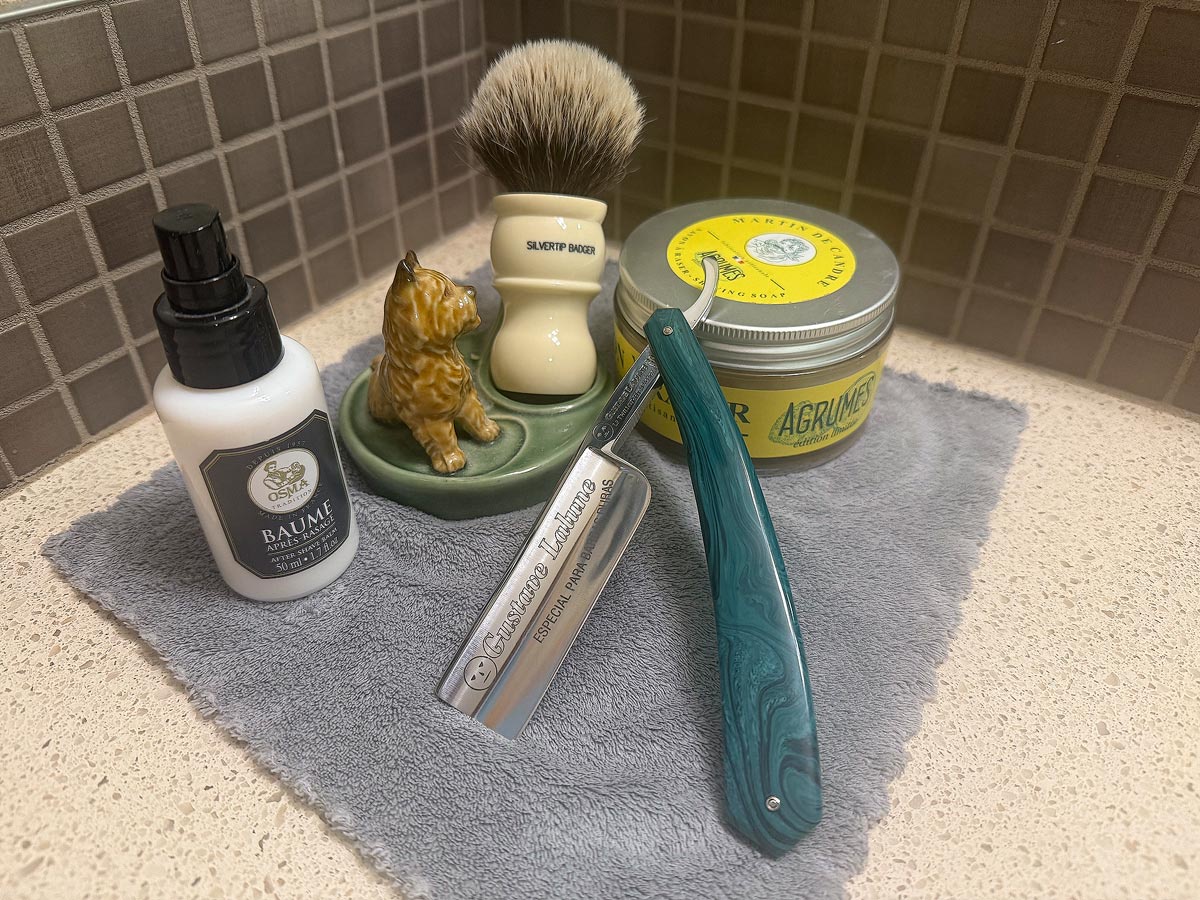 On a gray microfiber towel a straight razor with blade open about 45 degrees leans next to a bottle of aftershave and a jar of shave soap. A shave brush rests bristles-up in a green Wade ceramic pipe rest that features a brown Cairn Terrier.