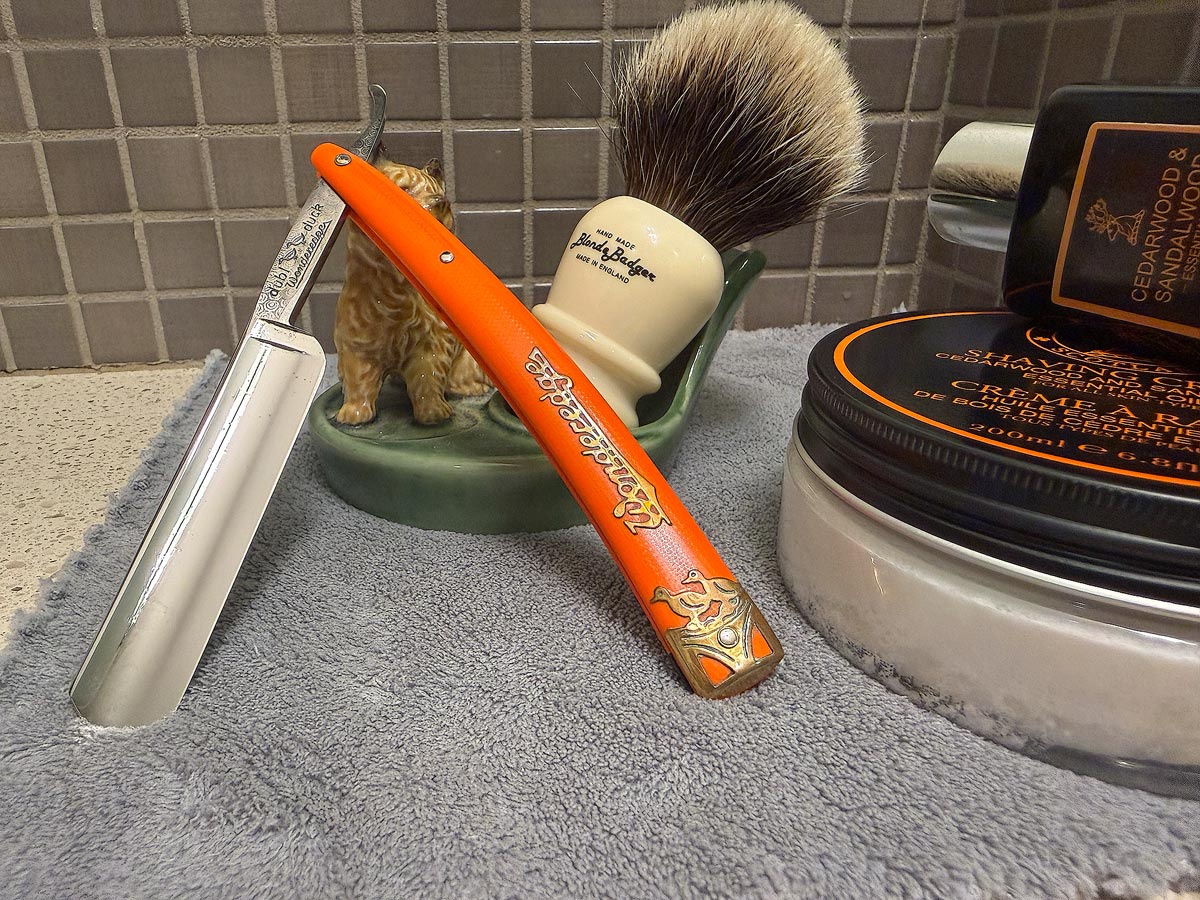 On a gray microfiber towel a straight razor with blade open about 45 degrees leans next to a bottle of aftershave and a jar of shave soap. A shave brush rests bristles-up in a green Wade ceramic pipe rest that features a brown Cairn Terrier.