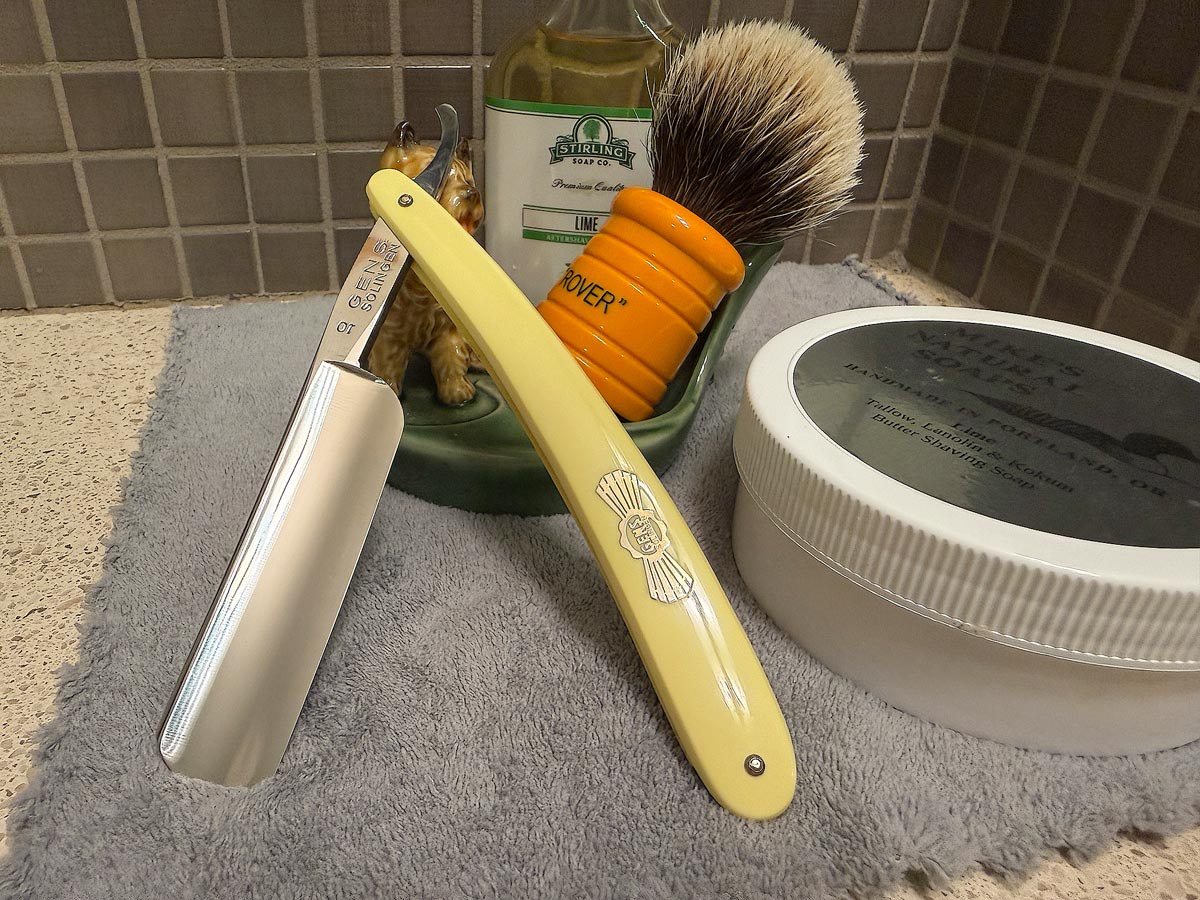 On a gray microfiber towel a straight razor with blade open about 45 degrees leans next to a bottle of aftershave and a jar of shave soap. A shave brush rests bristles-up in a green Wade ceramic pipe rest that features a brown Cairn Terrier.