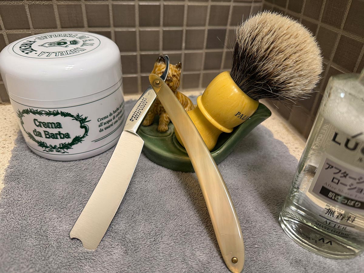 On a gray microfiber towel a straight razor with blade open about 45 degrees leans next to a bottle of aftershave and a jar of shave soap. A shave brush rests bristles-up in a green Wade ceramic pipe rest that features a brown Cairn Terrier.