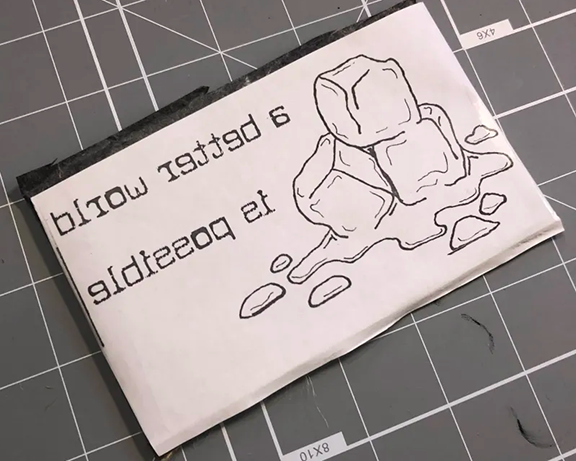 a reverse image of a melting stack of ice and the text "a better world is possible", being transferred to a linoleum block with some carbon paper