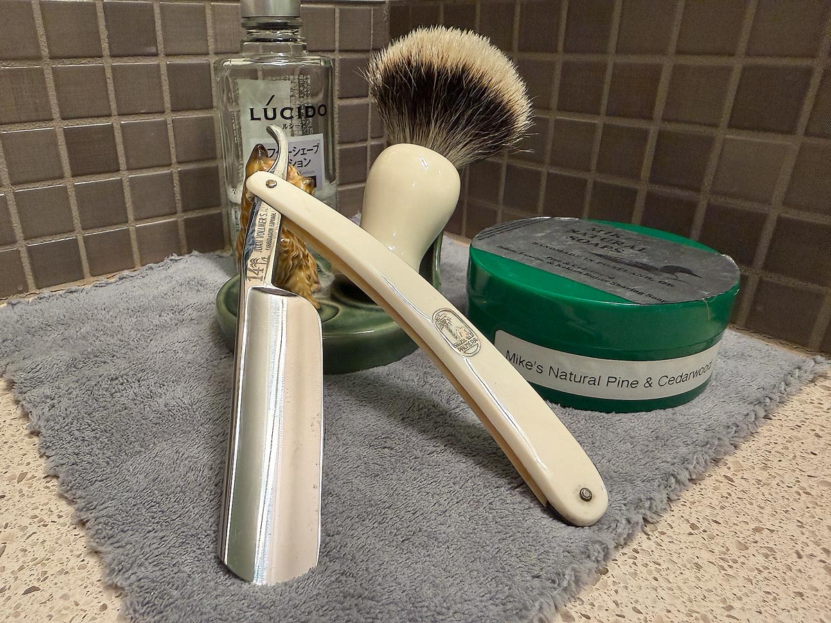 On a gray microfiber towel a straight razor with blade open about 45 degrees leans next to a bottle of aftershave and a jar of shave soap. A shave brush rests bristles-up in a green Wade ceramic pipe rest that features a brown Cairn Terrier.