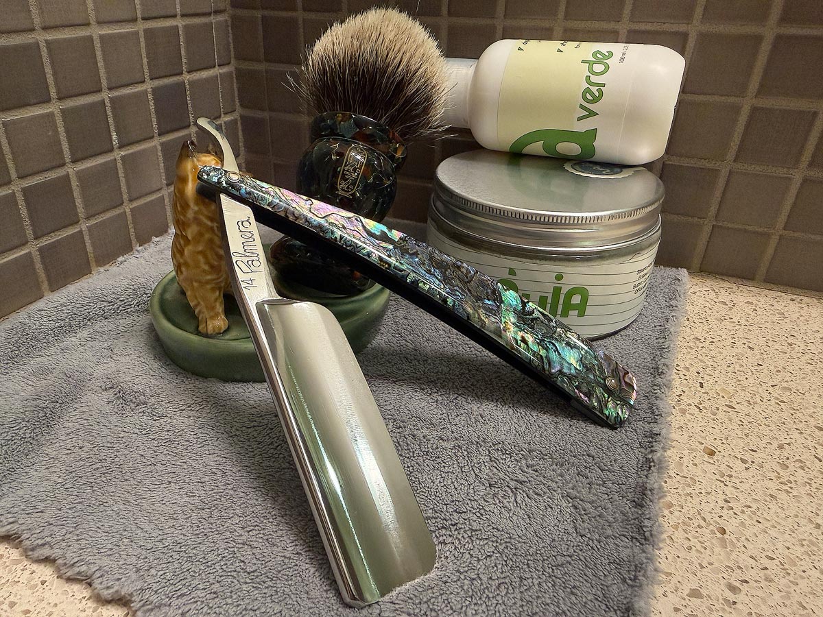 On a gray microfiber towel a straight razor with blade open about 45 degrees leans next to a bottle of aftershave and a jar of shave soap. A shave brush rests bristles-up in a green Wade ceramic pipe rest that features a brown Cairn Terrier.