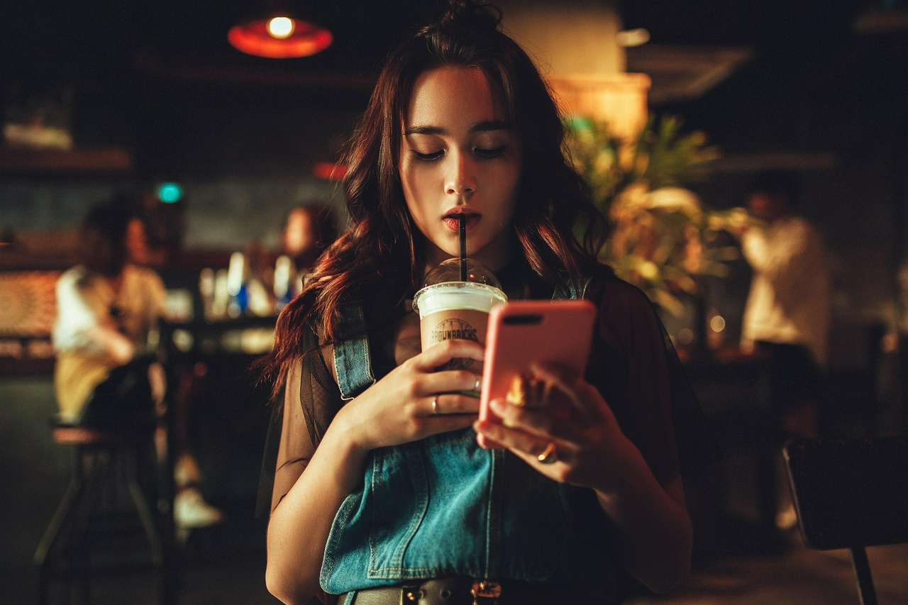 A teenage girl in a coffee shop looks at her phone while sipping a drink