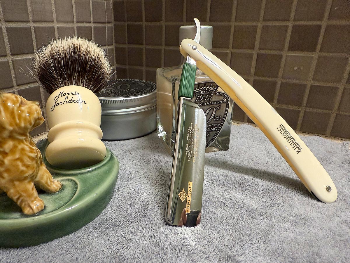On a gray microfiber towel a straight razor with blade open about 45 degrees leans next to a bottle of aftershave and a jar of shave soap. A shave brush rests bristles-up in a green Wade ceramic pipe rest that features a brown Cairn Terrier.