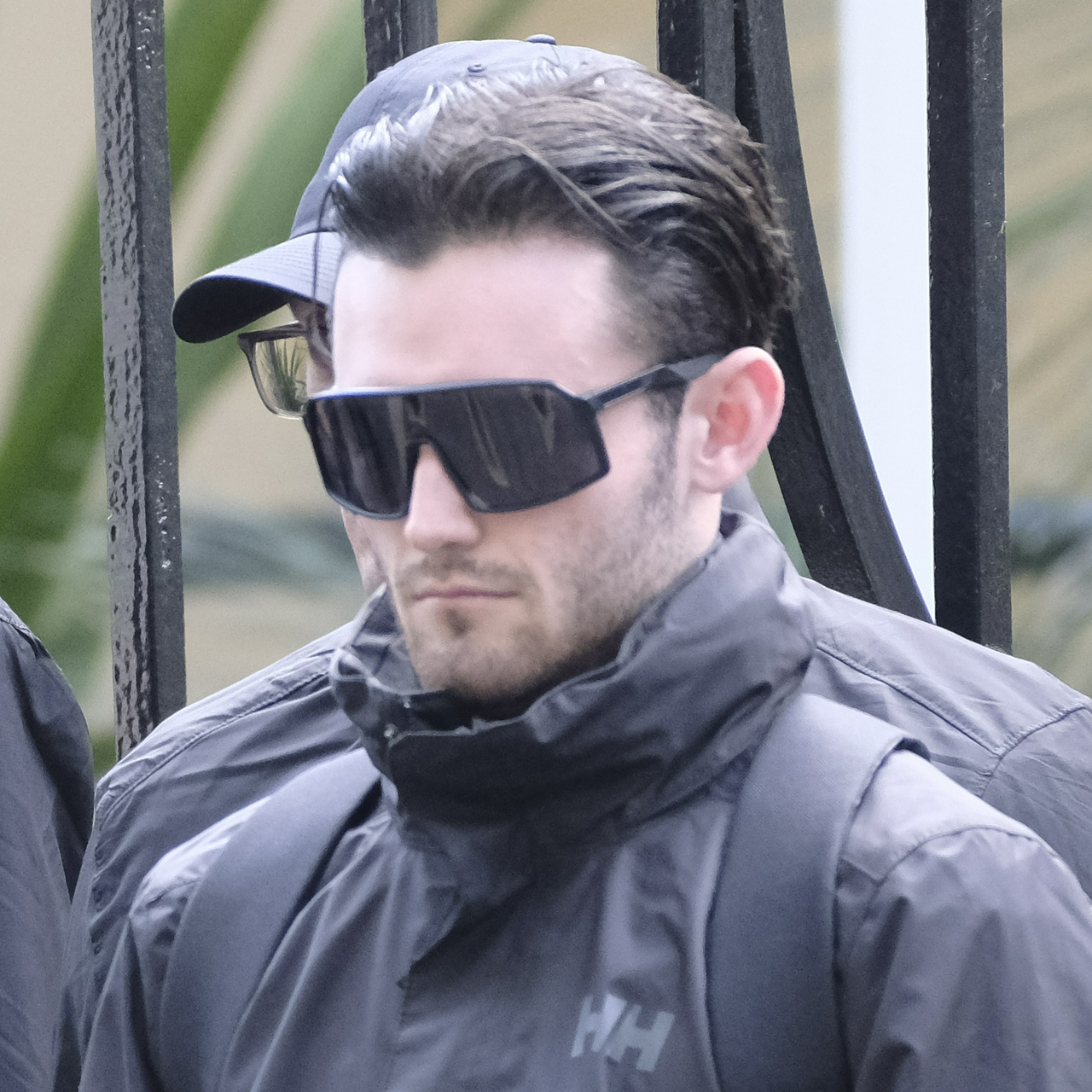 A scrawny man with a top-heavy head, round ears, facial scruff and huge black sunglasses