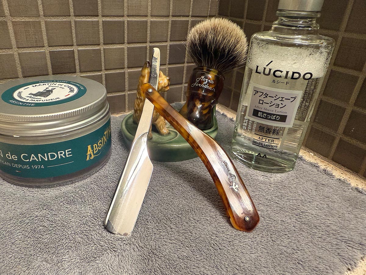 On a gray microfiber towel a straight razor with blade open about 45 degrees leans next to a bottle of aftershave and a jar of shave soap. A shave brush rests bristles-up in a green Wade ceramic pipe rest that features a brown Cairn Terrier.