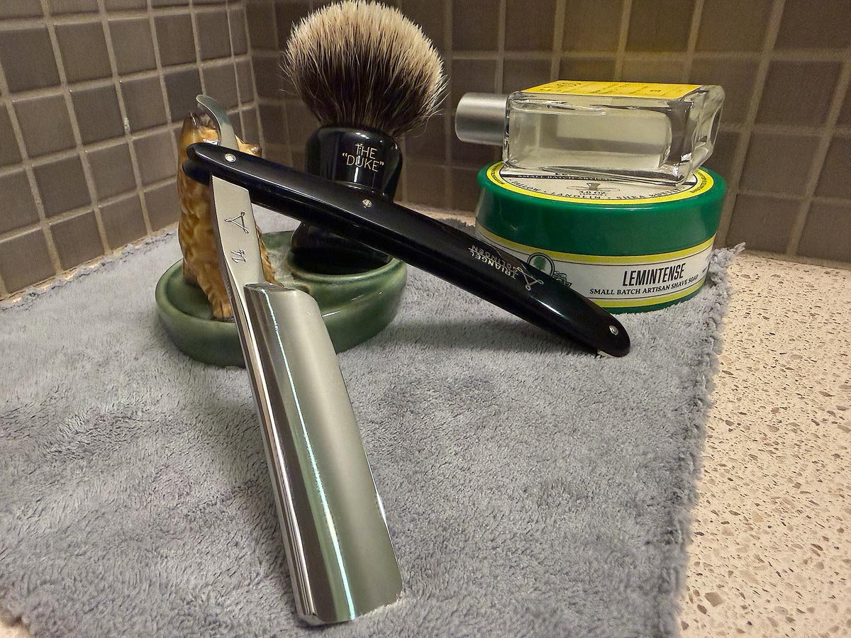 On a gray microfiber towel a straight razor with blade open about 45 degrees leans next to a bottle of aftershave and a jar of shave soap. A shave brush rests bristles-up in a green Wade ceramic pipe rest that features a brown Cairn Terrier.