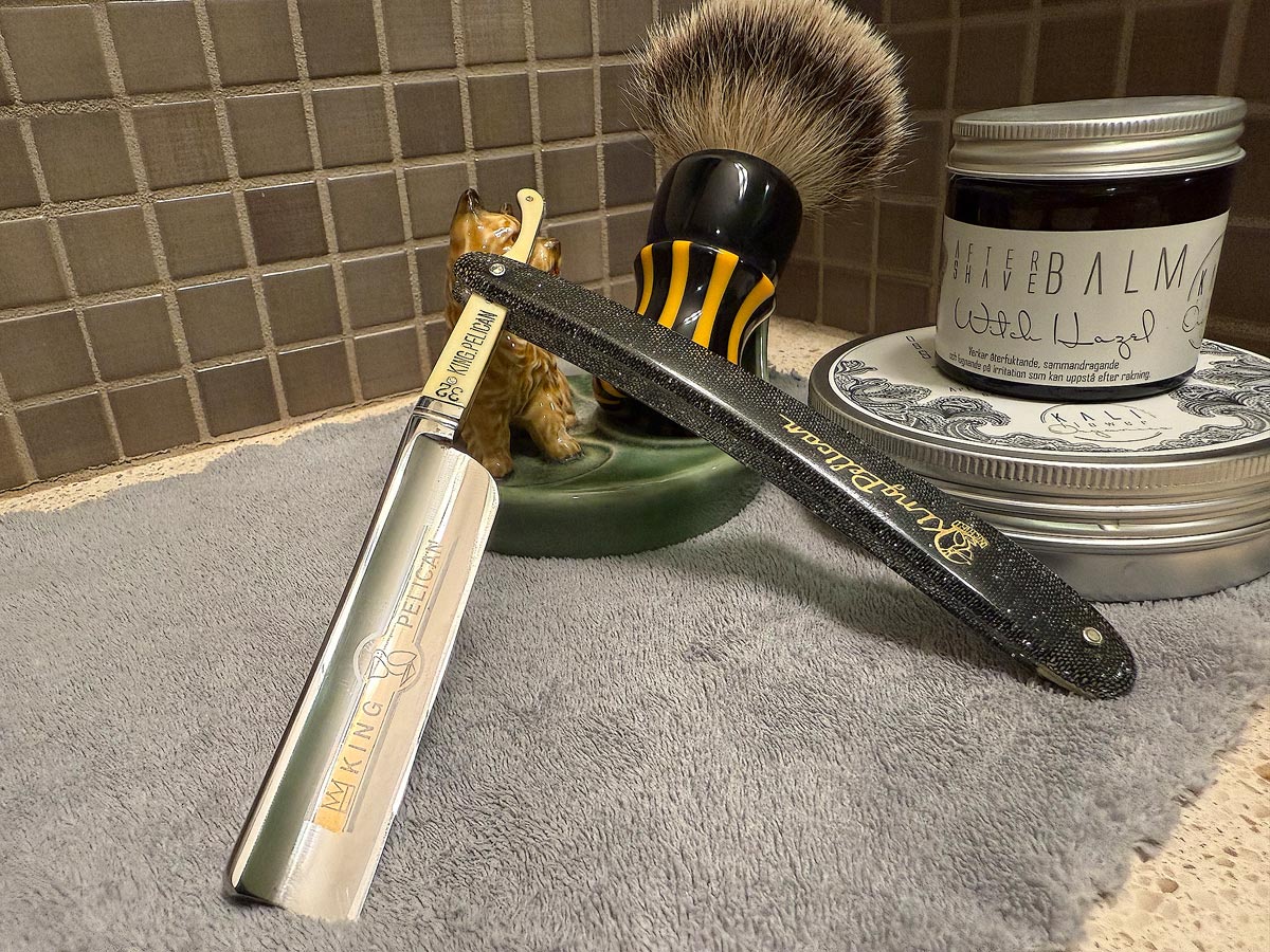 On a gray microfiber towel a straight razor with blade open about 45 degrees leans next to a bottle of aftershave and a jar of shave soap. A shave brush rests bristles-up in a green Wade ceramic pipe rest that features a brown Cairn Terrier.