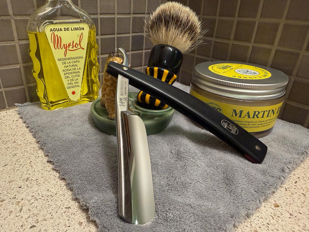 On a gray microfiber towel a straight razor with blade open about 45 degrees leans next to a bottle of aftershave and a jar of shave soap. A shave brush rests bristles-up in a green Wade ceramic pipe rest that features a brown Cairn Terrier.