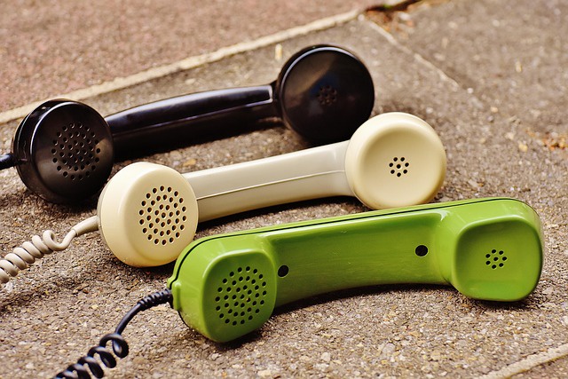 Three corded phone handsets laying on top of a hard surface like bricks or concrete blocks. Each handset has a cord connected to it. The top handset is black, the middle handset is cream colored. The bottom handset is a green color.