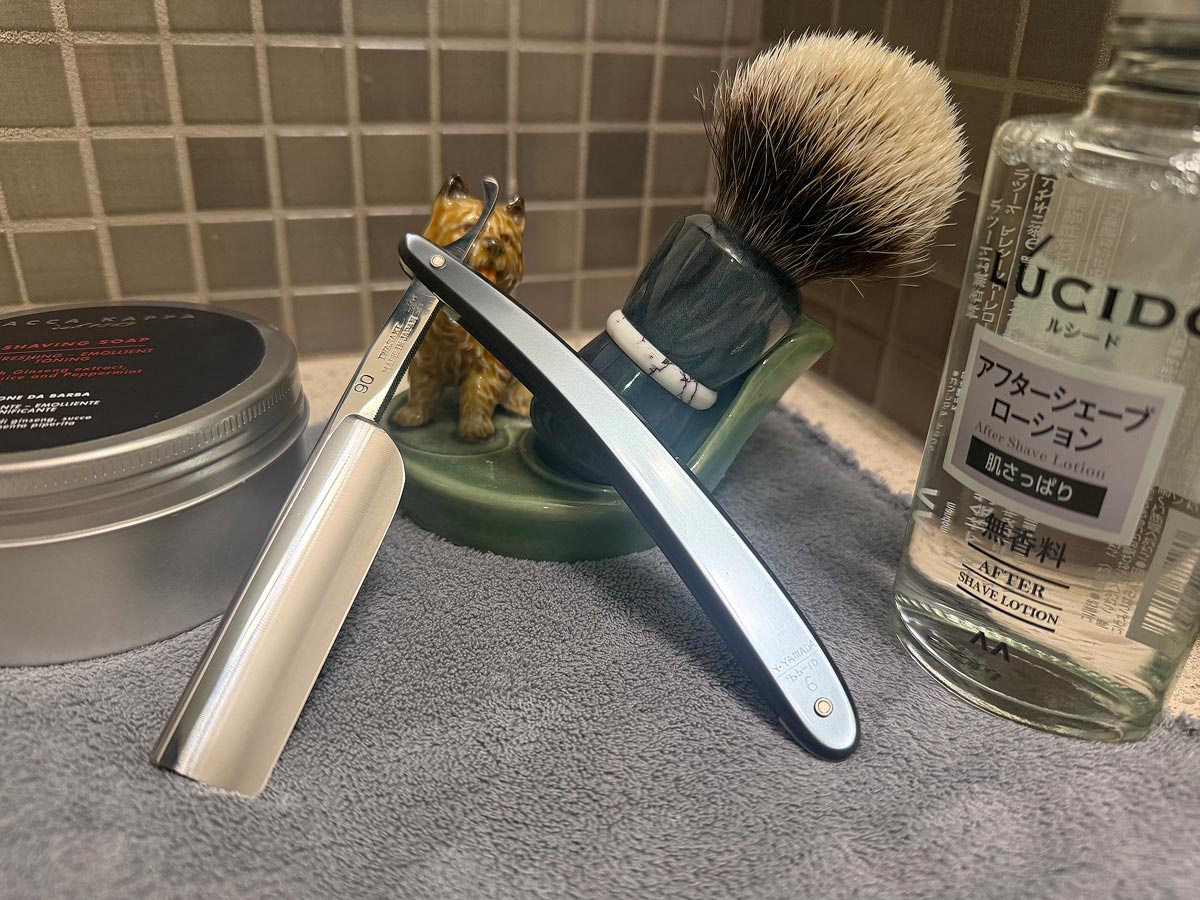 On a gray microfiber towel a straight razor with blade open about 45 degrees leans next to a bottle of aftershave and a jar of shave soap. A shave brush rests bristles-up in a green Wade ceramic pipe rest that features a brown Cairn Terrier.