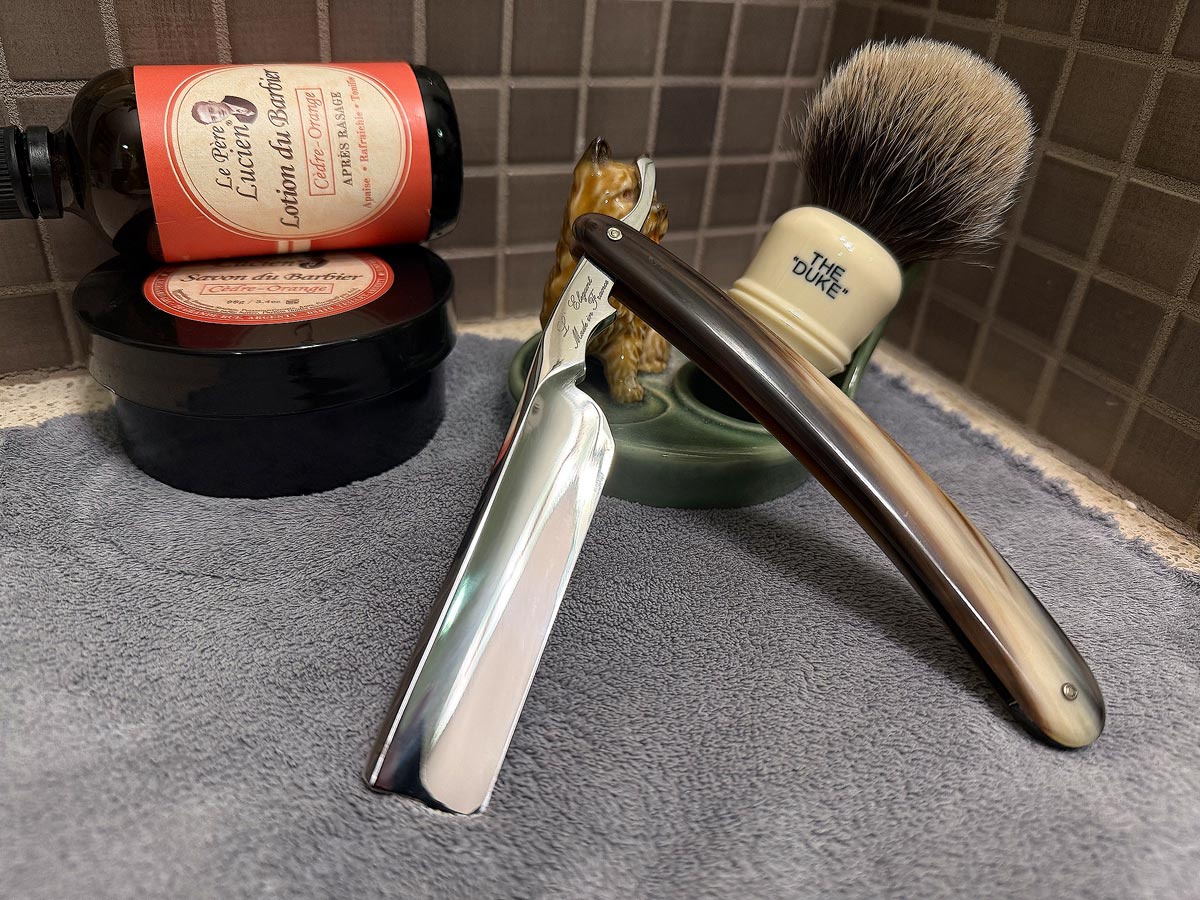 On a gray microfiber towel a straight razor with blade open about 45 degrees leans next to a bottle of aftershave and a jar of shave soap. A shave brush rests bristles-up in a green Wade ceramic pipe rest that features a brown Cairn Terrier.