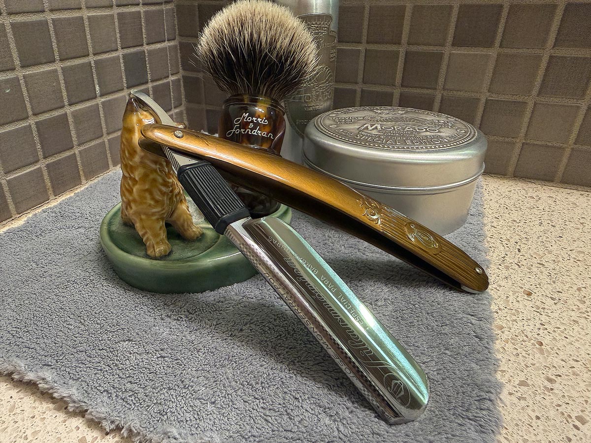 On a gray microfiber towel a straight razor with blade open about 45 degrees leans next to a bottle of aftershave and a jar of shave soap. A shave brush rests bristles-up in a green Wade ceramic pipe rest that features a brown Cairn Terrier.