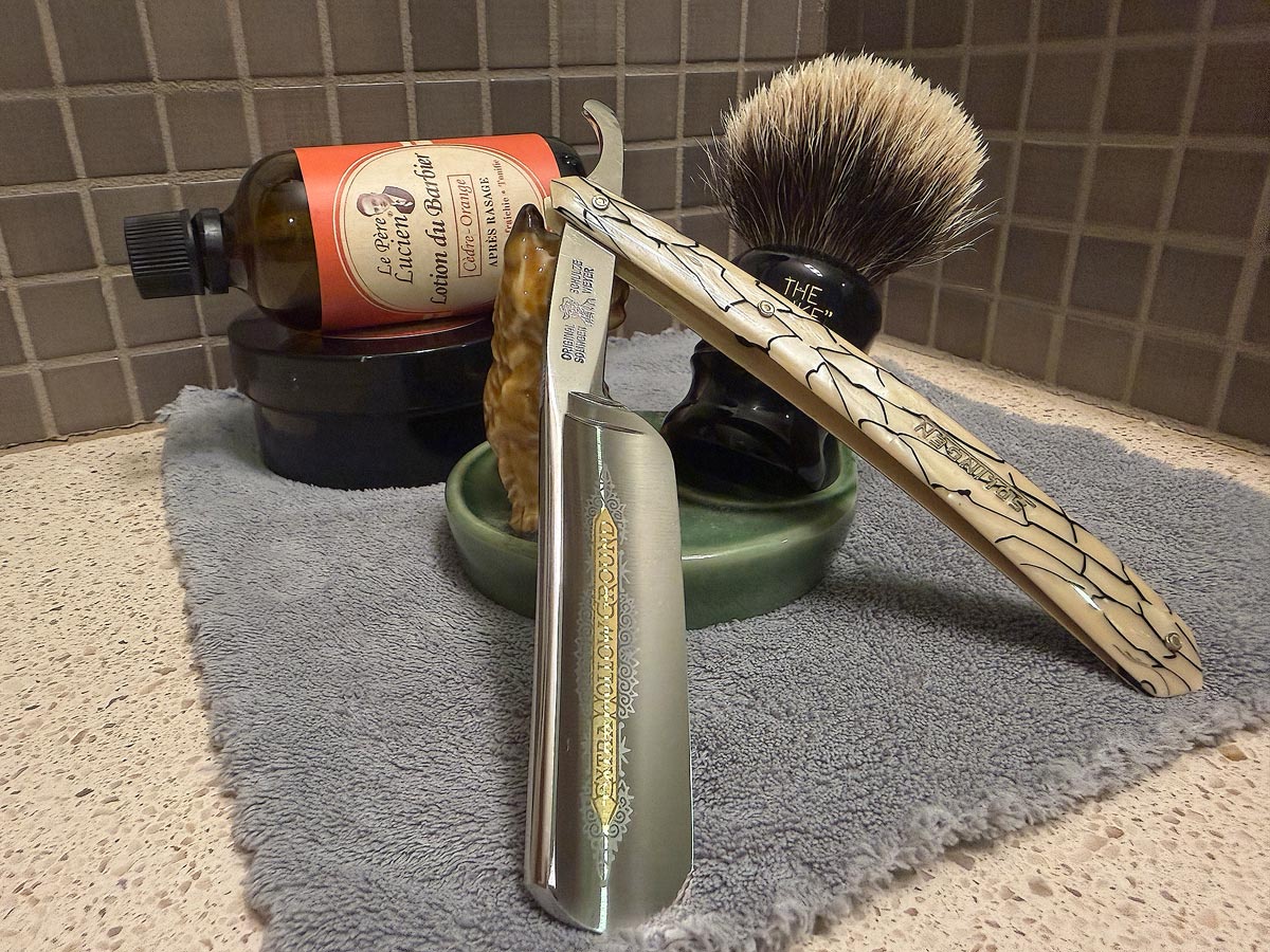 On a gray microfiber towel a straight razor with blade open about 45 degrees leans next to a bottle of aftershave and a jar of shave soap. A shave brush rests bristles-up in a green Wade ceramic pipe rest that features a brown Cairn Terrier.