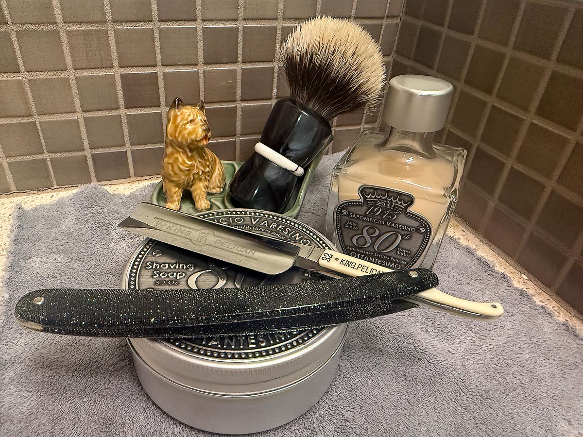 On a gray microfiber towel a straight razor with blade open about 45 degrees leans next to a bottle of aftershave and a jar of shave soap. A shave brush rests bristles-up in a green Wade ceramic pipe rest that features a brown Cairn Terrier.
