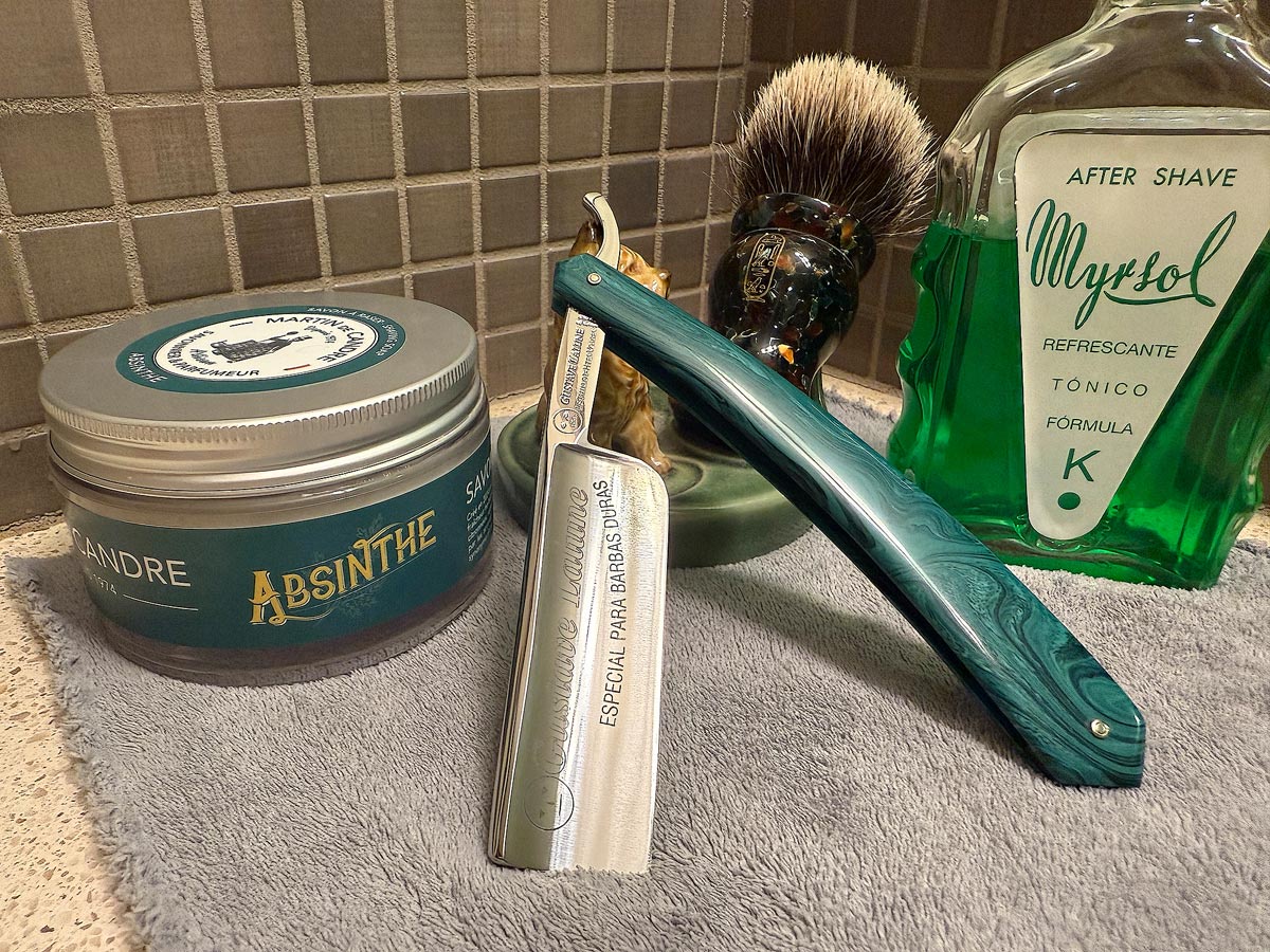 On a gray microfiber towel a straight razor with blade open about 45 degrees leans next to a bottle of aftershave and a jar of shave soap. A shave brush rests bristles-up in a green Wade ceramic pipe rest that features a brown Cairn Terrier.