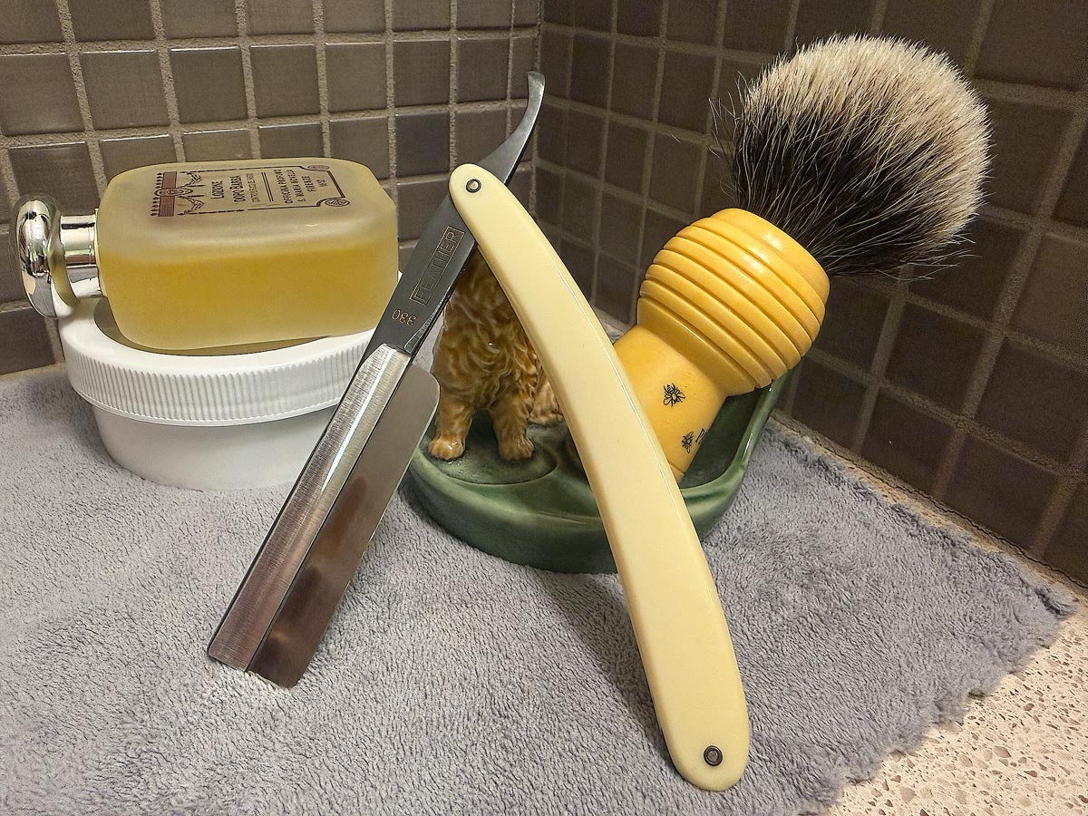 On a gray microfiber towel a straight razor with blade open about 45 degrees leans next to a bottle of aftershave and a jar of shave soap. A shave brush rests bristles-up in a green Wade ceramic pipe rest that features a brown Cairn Terrier.