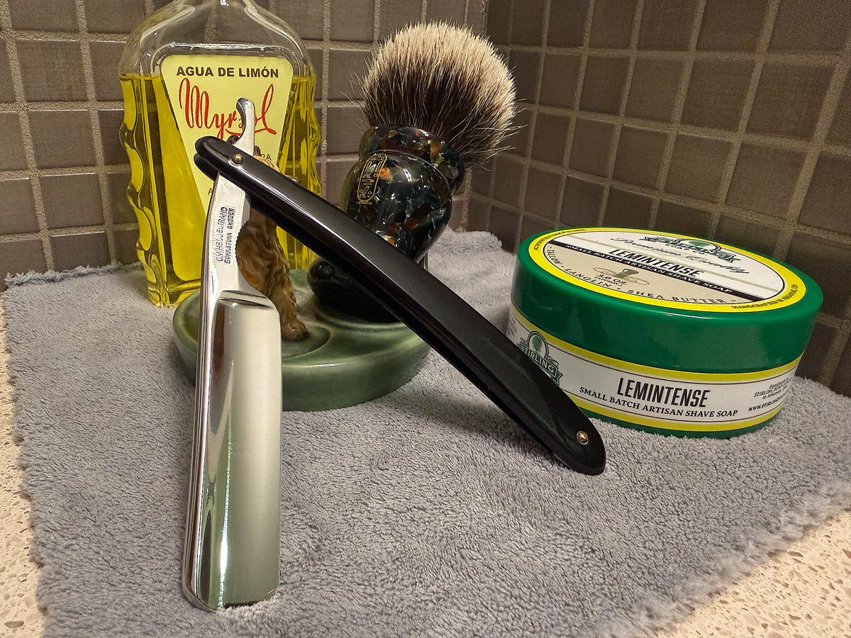 On a gray microfiber towel a straight razor with blade open about 45 degrees leans next to a bottle of aftershave and a jar of shave soap. A shave brush rests bristles-up in a green Wade ceramic pipe rest that features a brown Cairn Terrier.