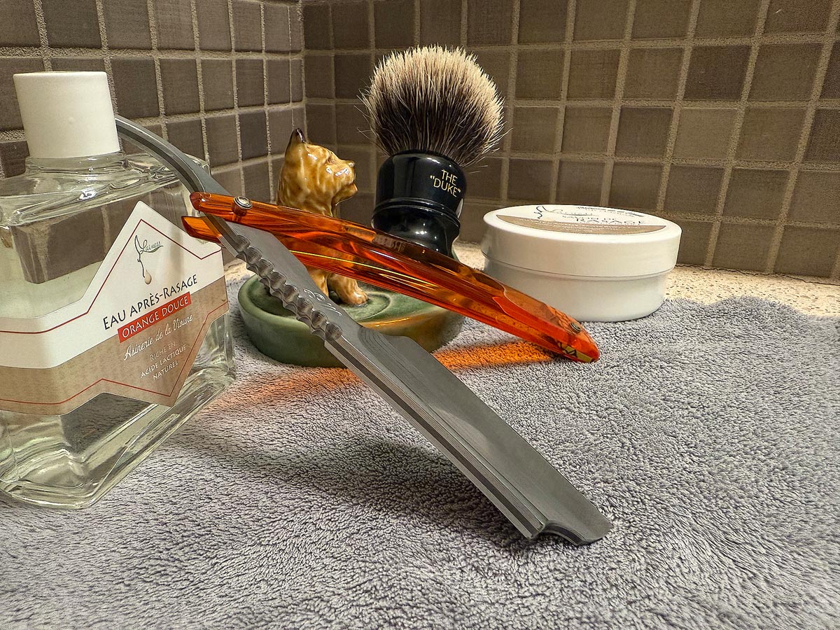 On a gray microfiber towel a straight razor with blade open about 45 degrees leans next to a bottle of aftershave and a jar of shave soap. A shave brush rests bristles-up in a green Wade ceramic pipe rest that features a brown Cairn Terrier.
