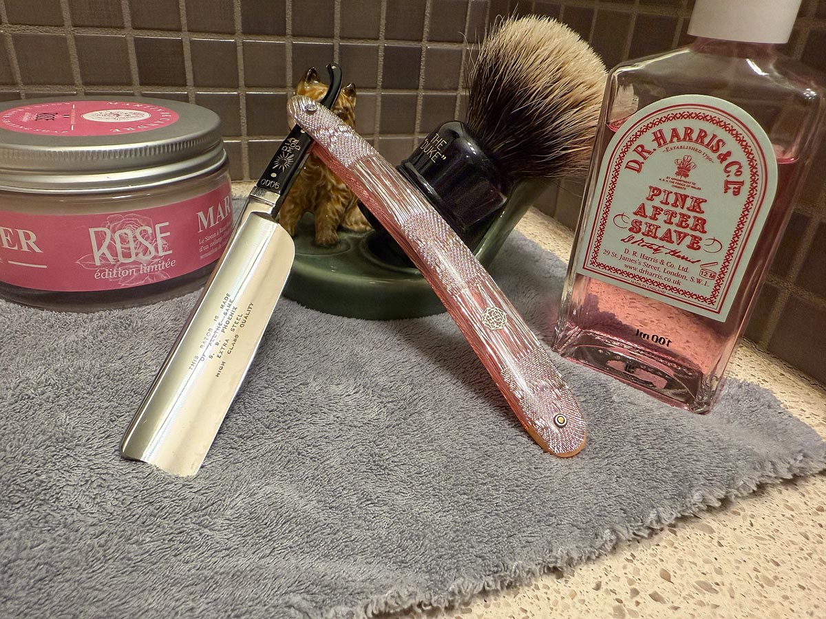 On a gray microfiber towel a straight razor with blade open about 45 degrees leans next to a bottle of aftershave and a jar of shave soap. A shave brush rests bristles-up in a green Wade ceramic pipe rest that features a brown Cairn Terrier.