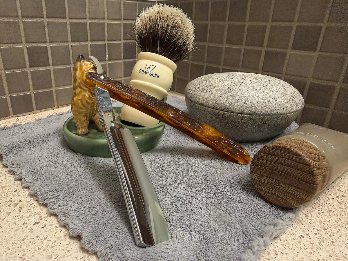 On a gray microfiber towel a straight razor with blade open about 45 degrees leans next to a bottle of aftershave and a jar of shave soap. A shave brush rests bristles-up in a green Wade ceramic pipe rest that features a brown Cairn Terrier.