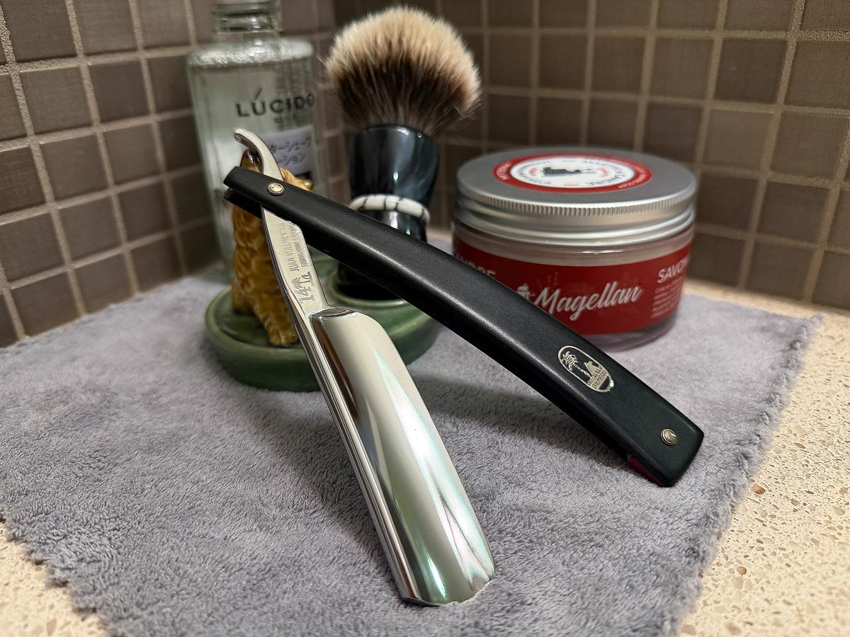 On a gray microfiber towel a straight razor with blade open about 45 degrees leans next to a bottle of aftershave and a jar of shave soap. A shave brush rests bristles-up in a green Wade ceramic pipe rest that features a brown Cairn Terrier.