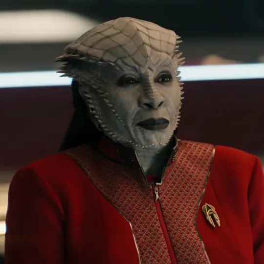 Gina Yashere as the Star Trek character Lura Thok. She wears a read starfleet uniform with her collar unzipped partially.