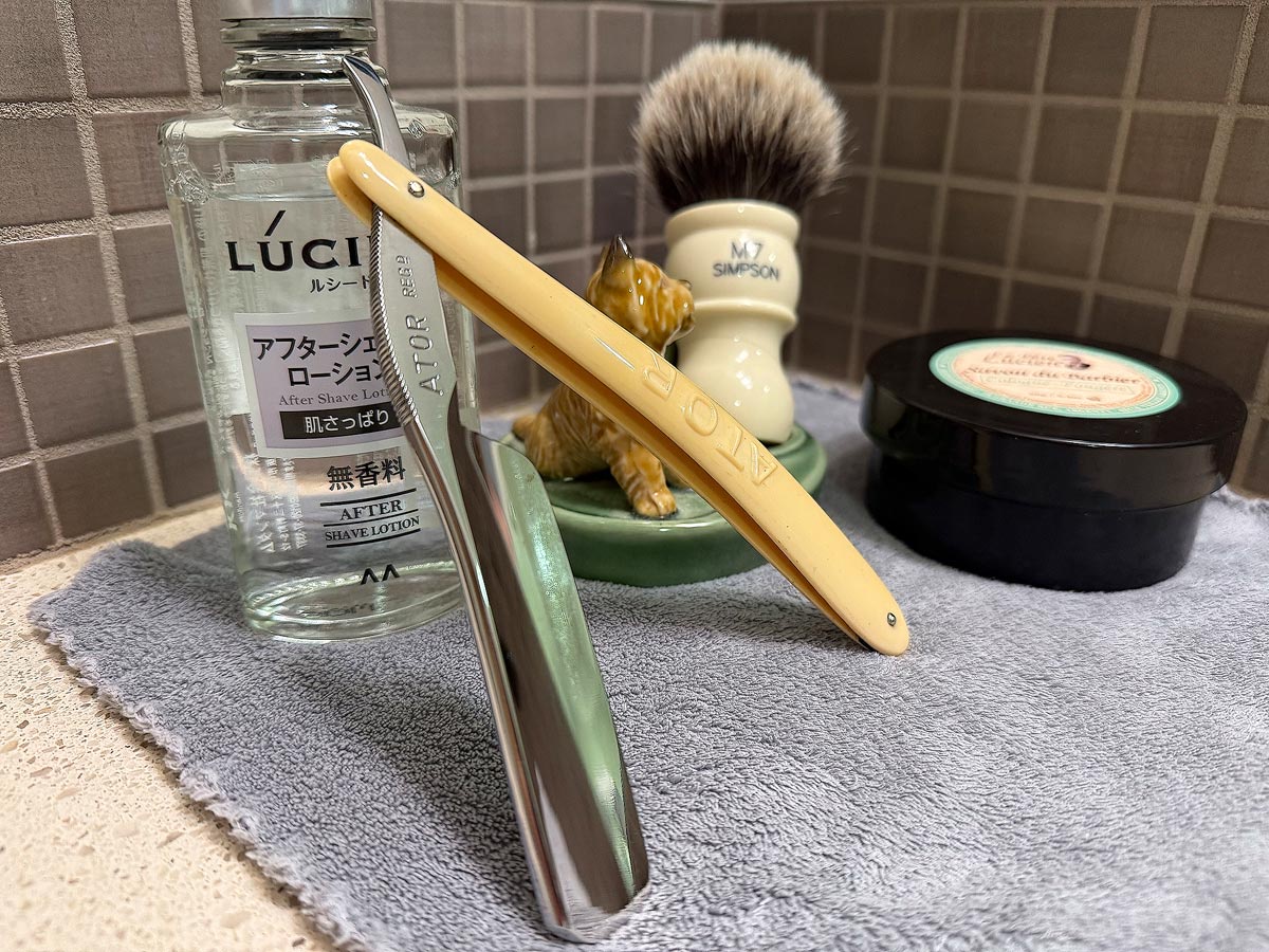 On a gray microfiber towel a straight razor with blade open about 45 degrees leans next to a bottle of aftershave and a jar of shave soap. A shave brush rests bristles-up in a green Wade ceramic pipe rest that features a brown Cairn Terrier.