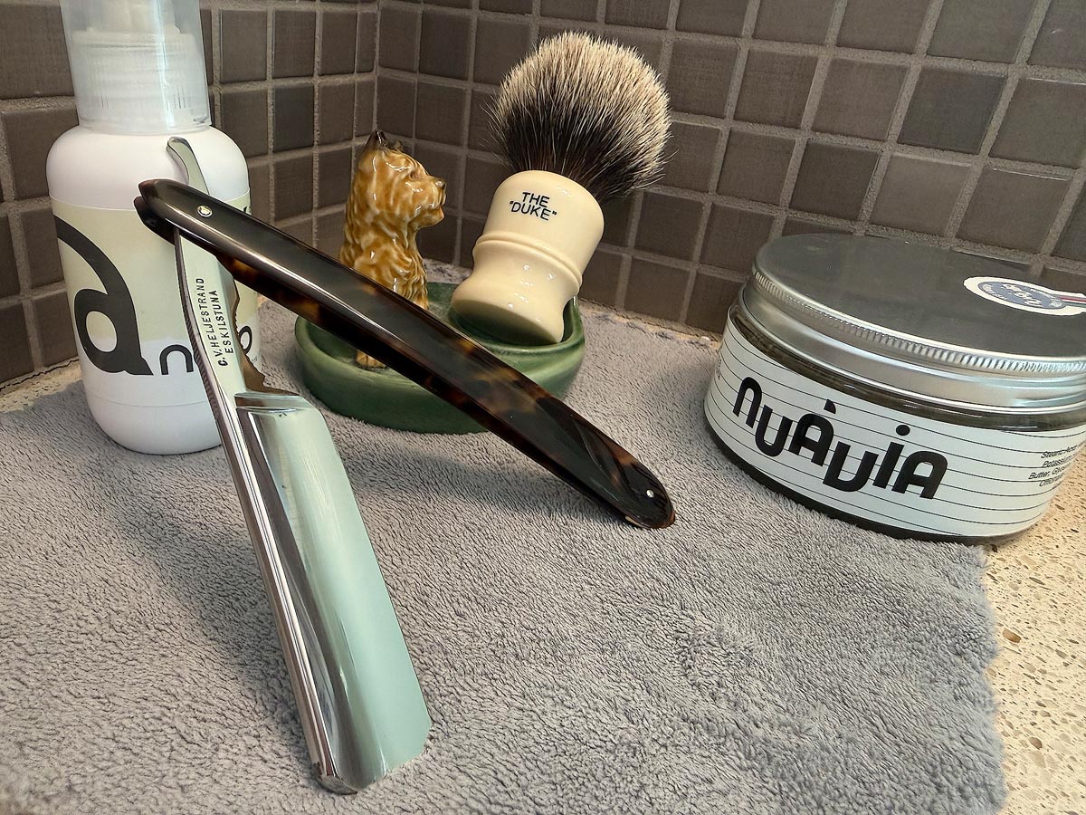 On a gray microfiber towel a straight razor with blade open about 45 degrees leans next to a bottle of aftershave and a jar of shave soap. A shave brush rests bristles-up in a green Wade ceramic pipe rest that features a brown Cairn Terrier.