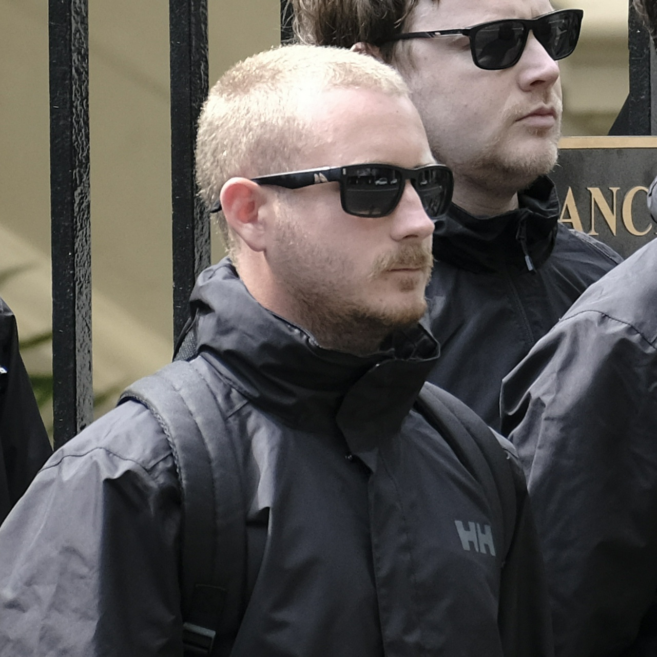 A short scruffy blonde man with a small, stern mouth and overgrown whiskers with sunglasses on