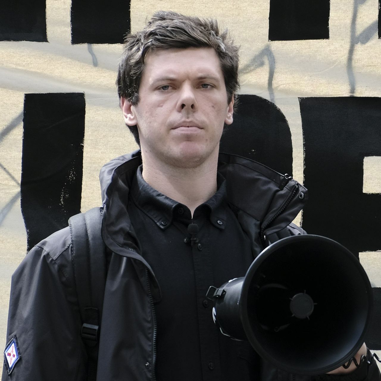 Eltis's pockmarked sad face looks down the lens, he's holding Joel's megaphone and has a lapel mic on