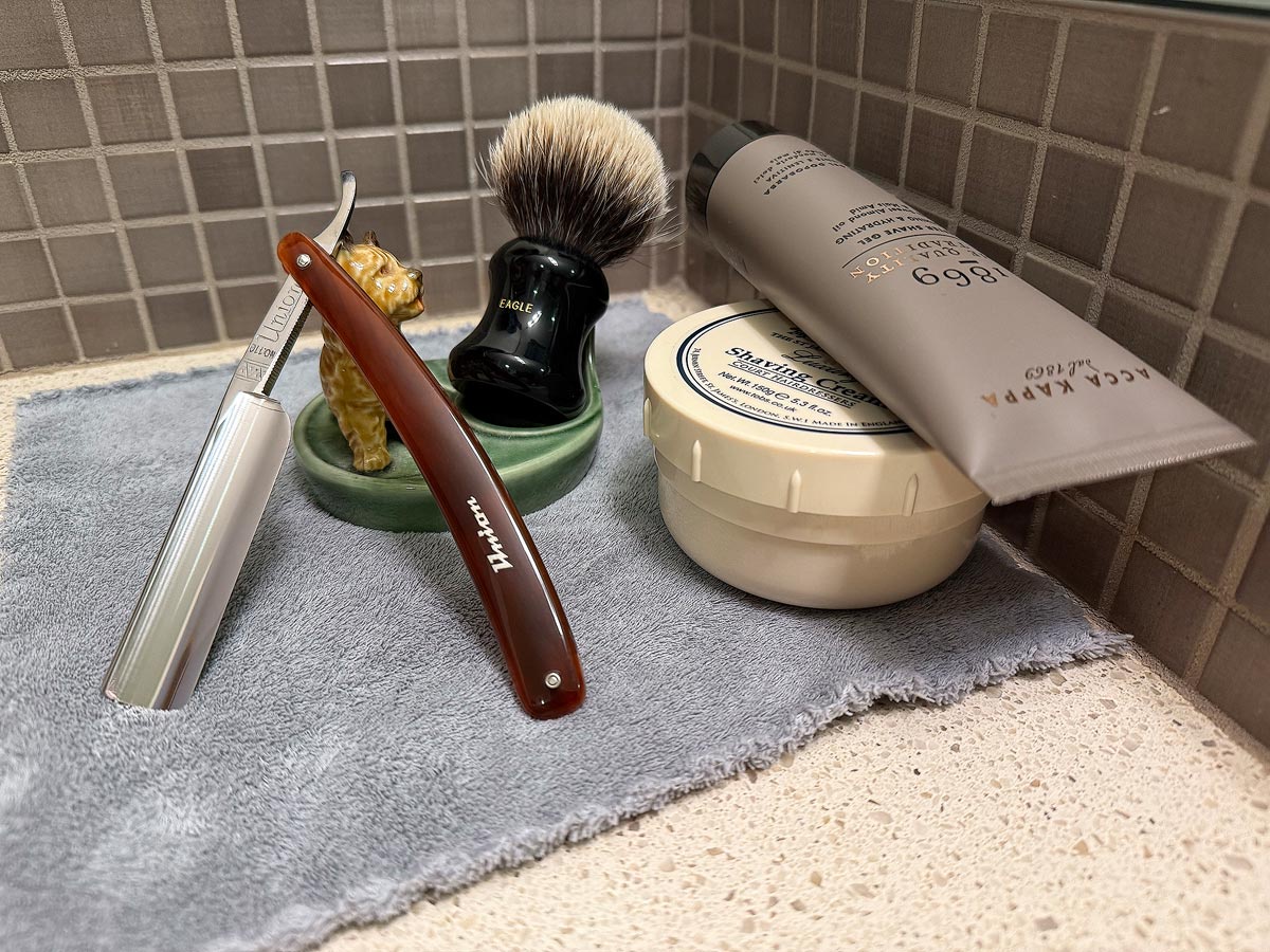 On a gray microfiber towel a straight razor with blade open about 45 degrees leans next to a bottle of aftershave and a jar of shave soap. A shave brush rests bristles-up in a green Wade ceramic pipe rest that features a brown Cairn Terrier.