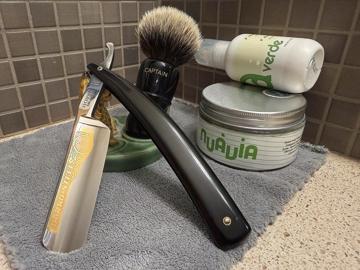 On a gray microfiber towel a straight razor with blade open about 45 degrees leans next to a bottle of aftershave and a jar of shave soap. A shave brush rests bristles-up in a green Wade ceramic pipe rest that features a brown Cairn Terrier.