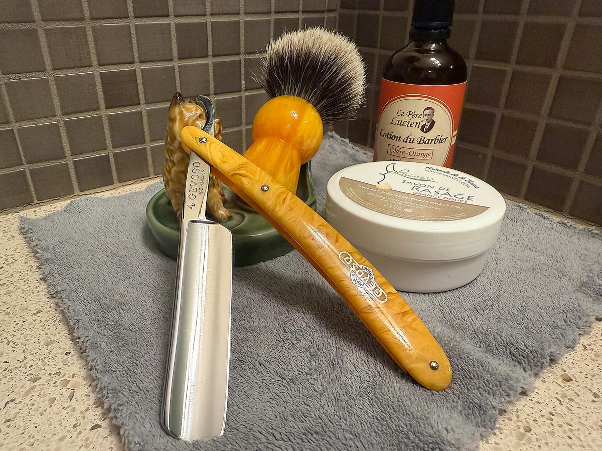 On a gray microfiber towel a straight razor with blade open about 45 degrees leans next to a bottle of aftershave and a jar of shave soap. A shave brush rests bristles-up in a green Wade ceramic pipe rest that features a brown Cairn Terrier.