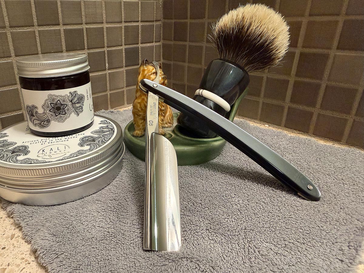 On a gray microfiber towel a straight razor with blade open about 45 degrees leans next to a bottle of aftershave and a jar of shave soap. A shave brush rests bristles-up in a green Wade ceramic pipe rest that features a brown Cairn Terrier.