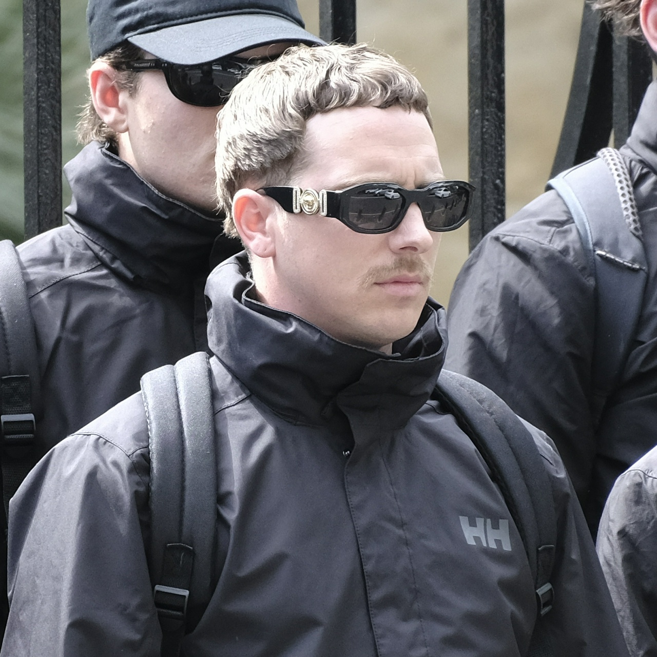 A shorter guy with short, carefully arranged curls and designer-look sunglasses with a gaudy gold motif on the arm, and a sparse moustache that bristles as he frowns