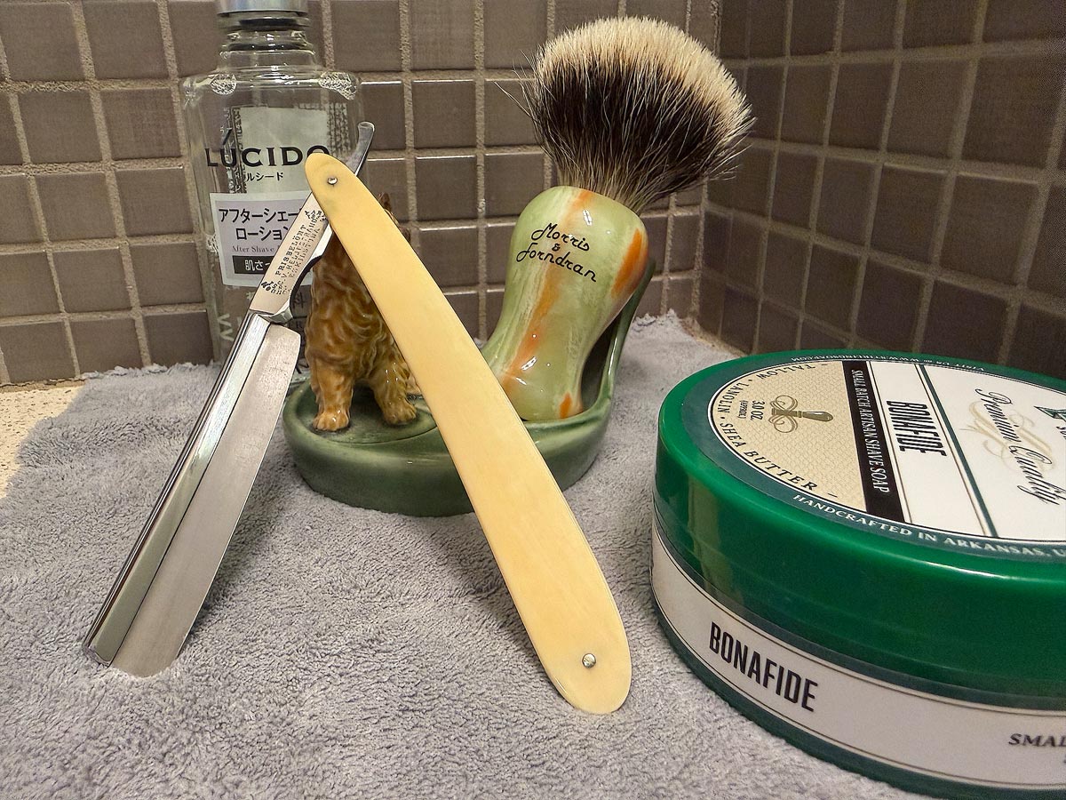 On a gray microfiber towel a straight razor with blade open about 45 degrees leans next to a bottle of aftershave and a jar of shave soap. A shave brush rests bristles-up in a green Wade ceramic pipe rest that features a brown Cairn Terrier.