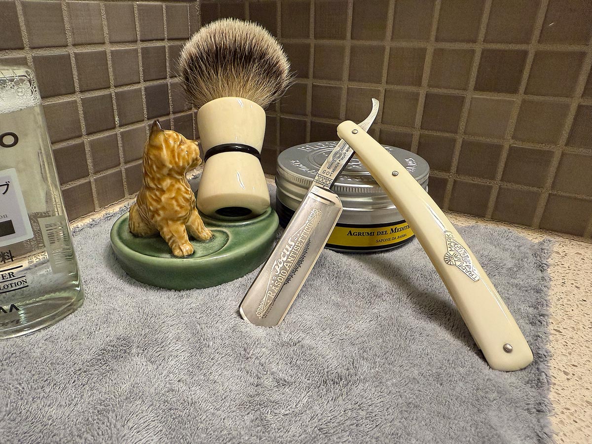 On a gray microfiber towel a straight razor with blade open about 45 degrees leans next to a bottle of aftershave and a jar of shave soap. A shave brush rests bristles-up in a green Wade ceramic pipe rest that features a brown Cairn Terrier.