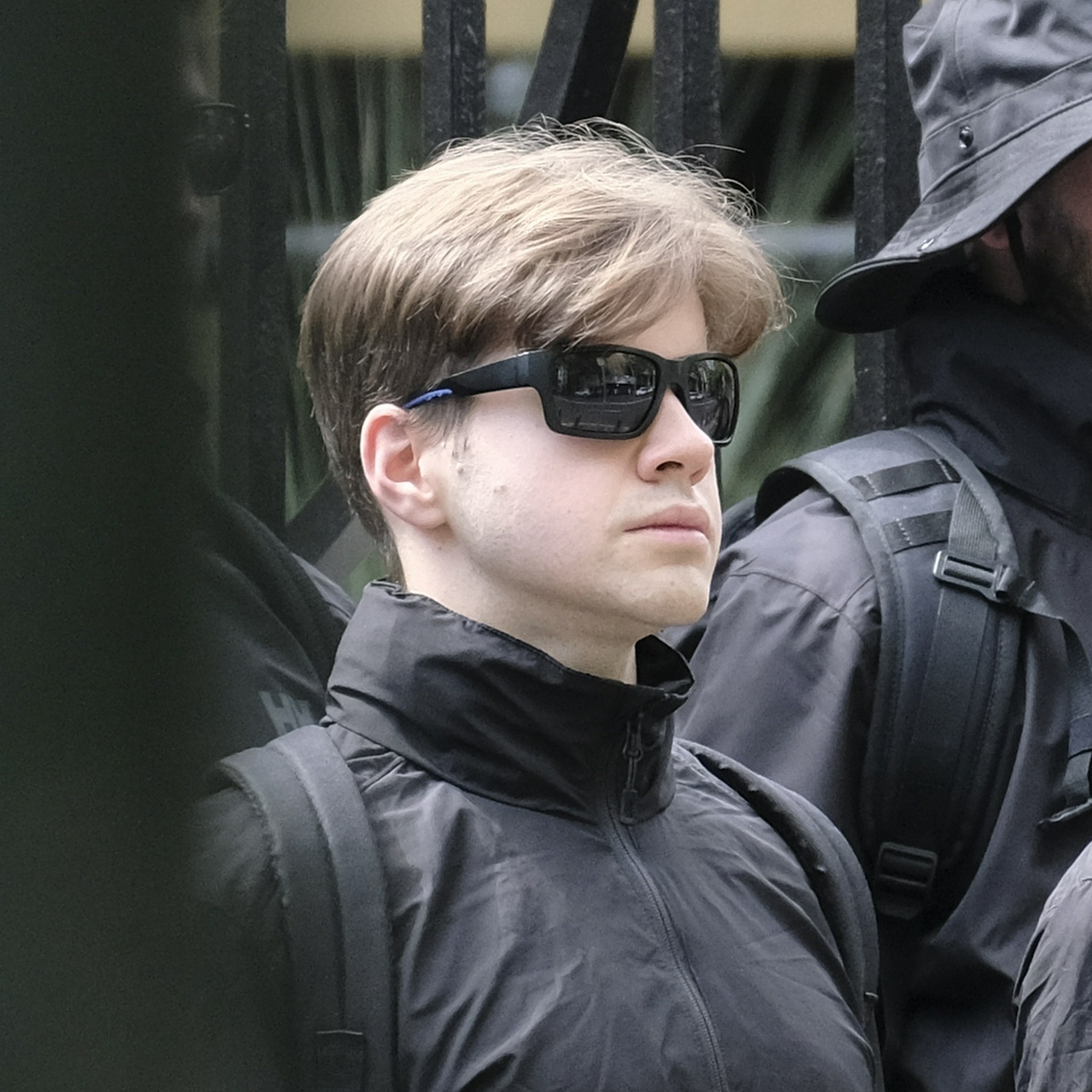 A young white man with rounded features and straight brown hair wearing black sunglasses