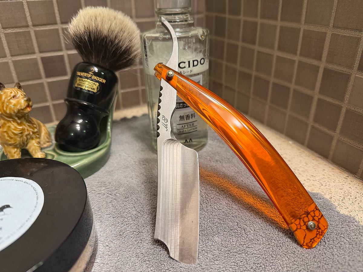 On a gray microfiber towel a straight razor with blade open about 45 degrees leans next to a bottle of aftershave and a jar of shave soap. A shave brush rests bristles-up in a green Wade ceramic pipe rest that features a brown Cairn Terrier.